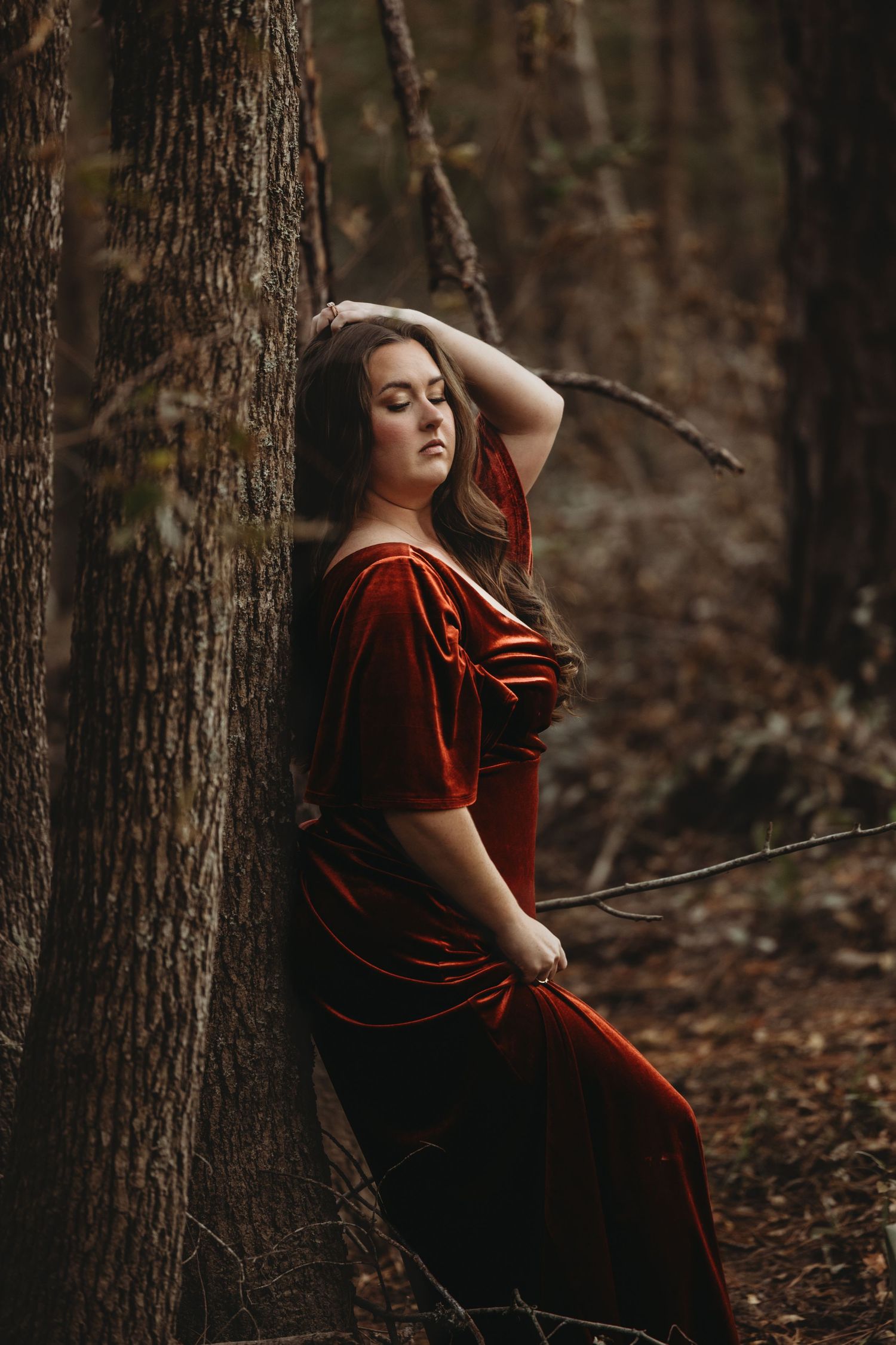 A moody portrait of a person in a red velvet dress leaning against a tree in the forest.