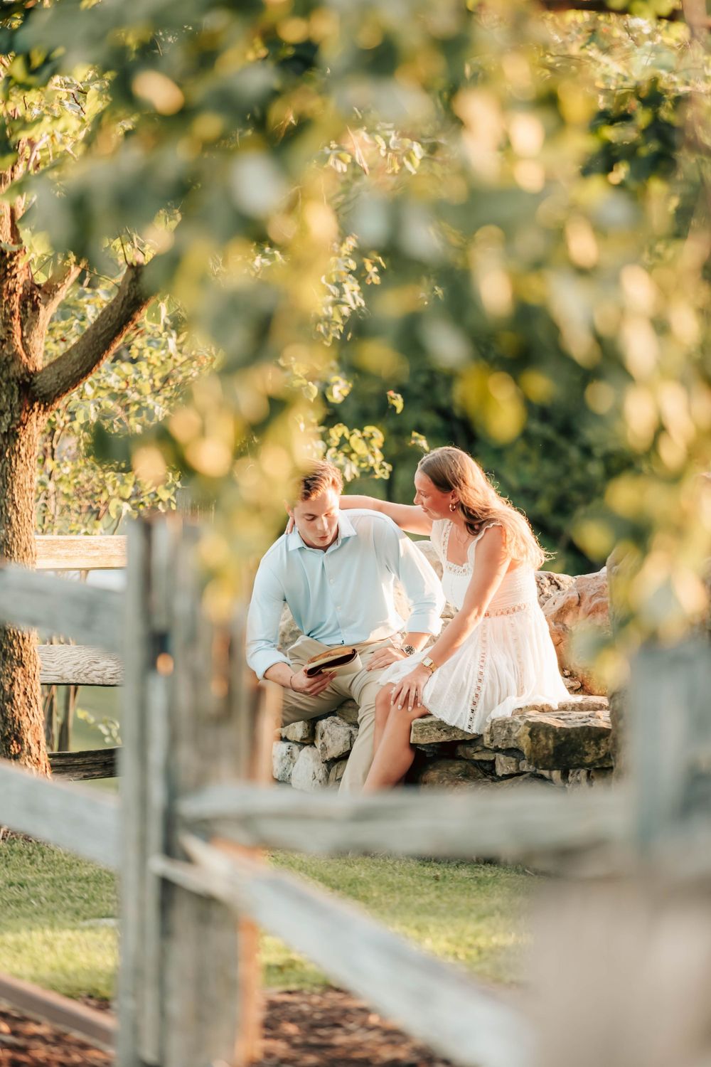 A couple sits together on a rustic stone bench during a romantic outdoor photoshoot at sunset with bokeh lighting.