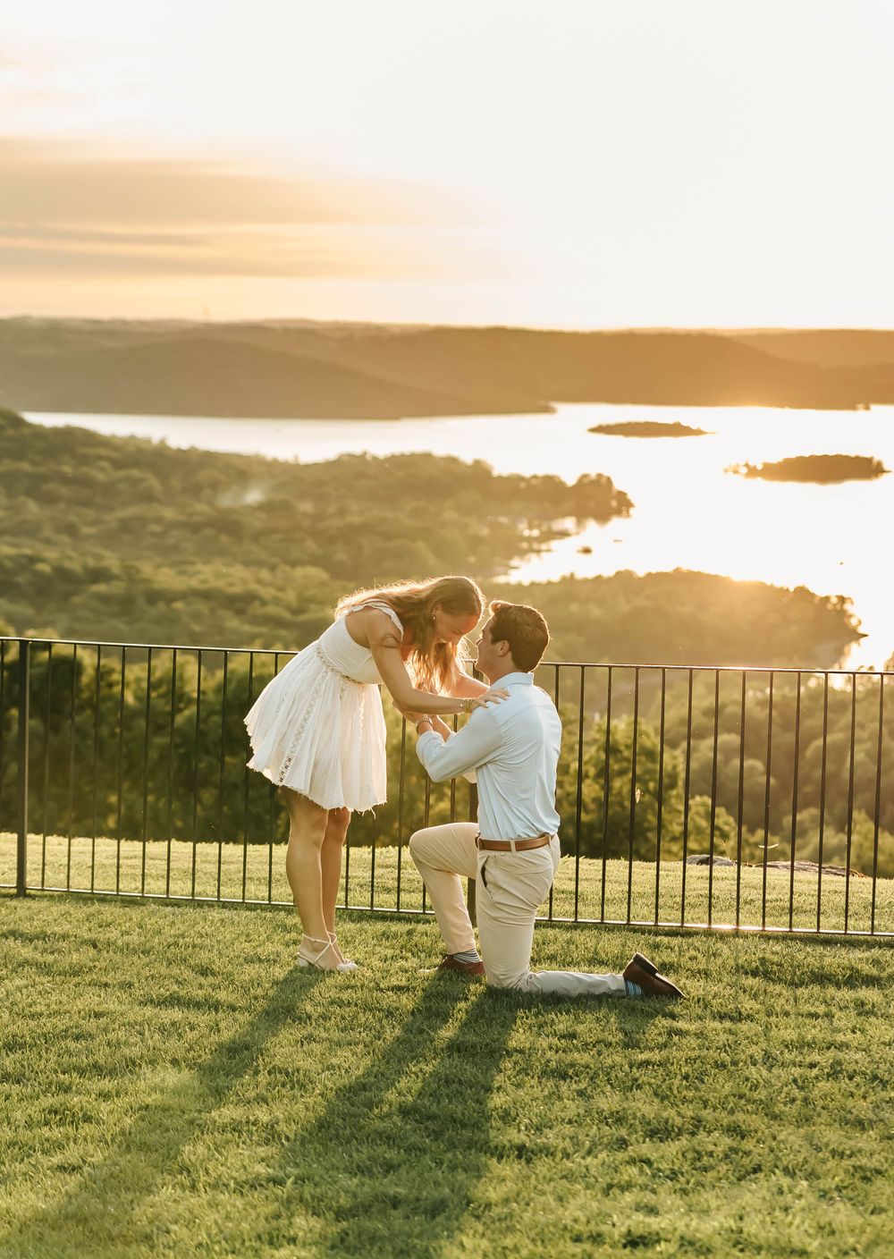A couple shares a romantic moment at sunset on a hilltop overlooking a scenic bay with water and islands in the distance.