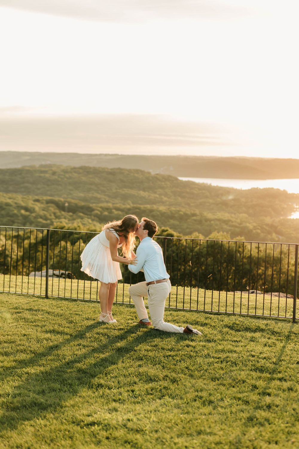 A romantic proposal moment at sunset overlooking a scenic coastal vista with rolling hills and an iron fence.