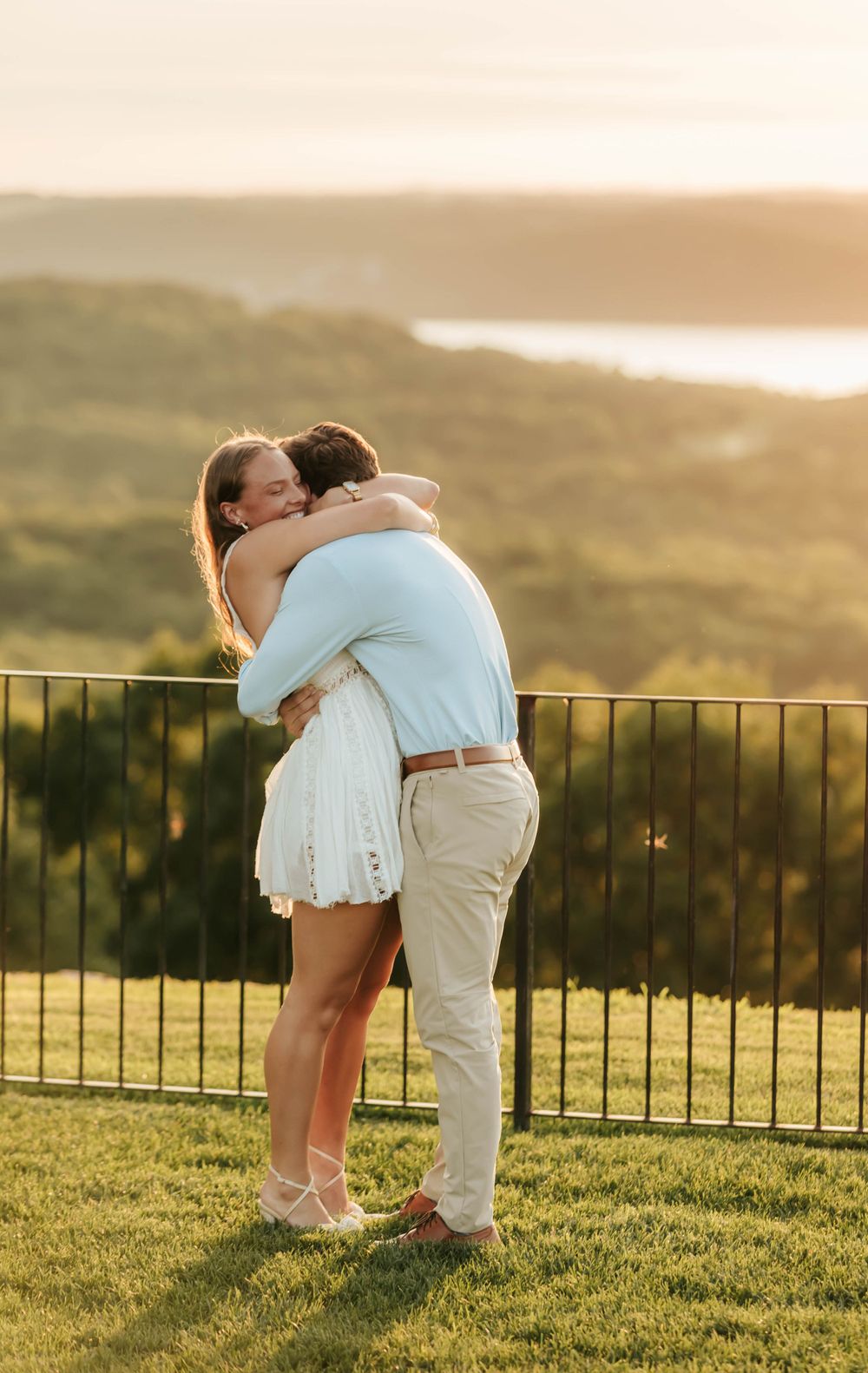 A couple embraces in a warm sunset on a hilltop overlook with a metal railing and grassy foreground.