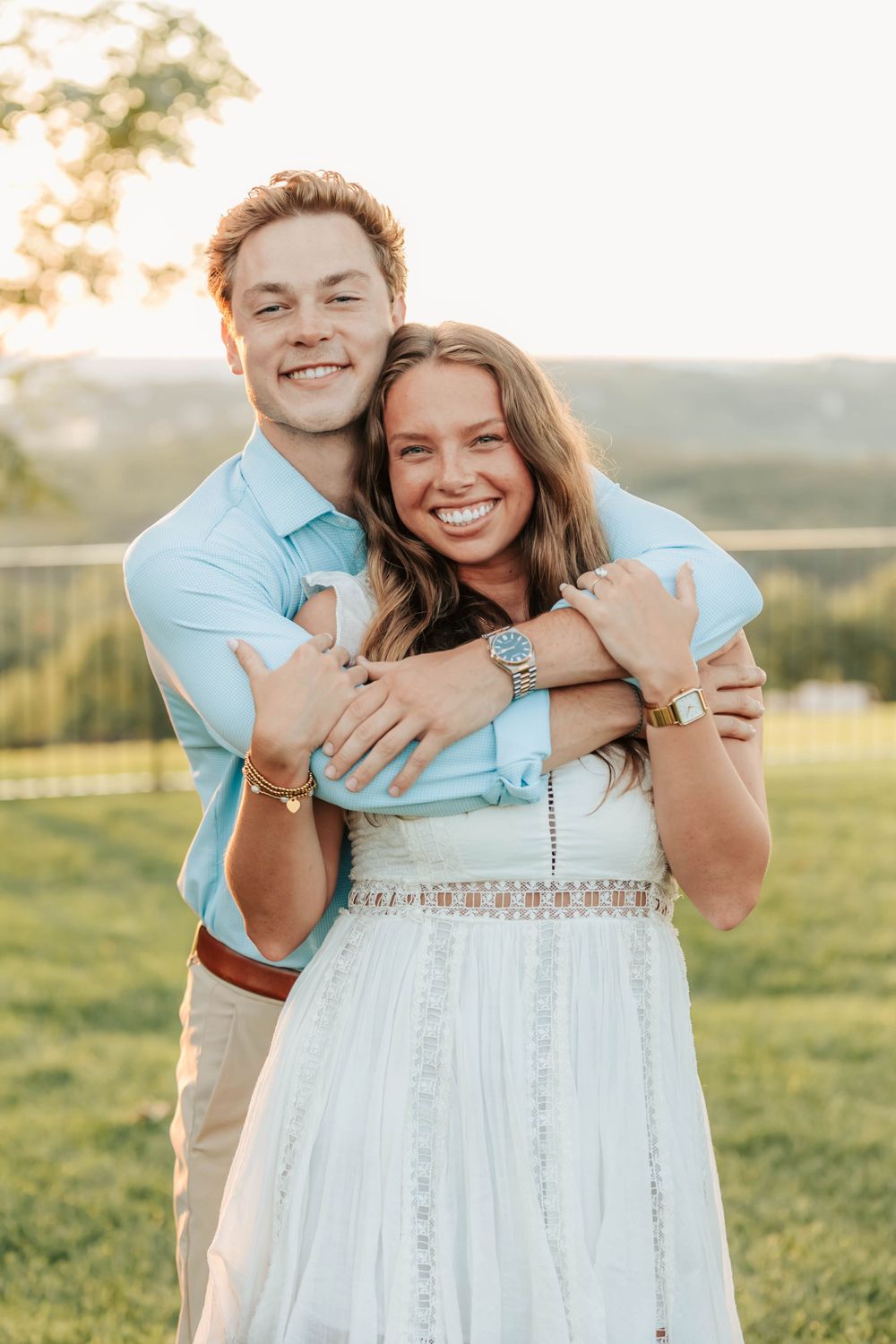 A happy couple embraces outdoors at sunset in a grassy field, the woman wearing a white dress and man in light blue shirt.