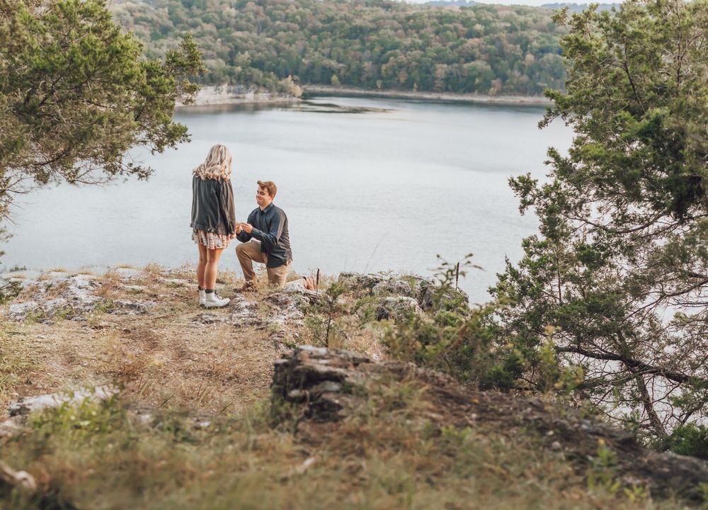 A scenic marriage proposal moment on a rocky cliff overlooking a serene lake surrounded by green forest.