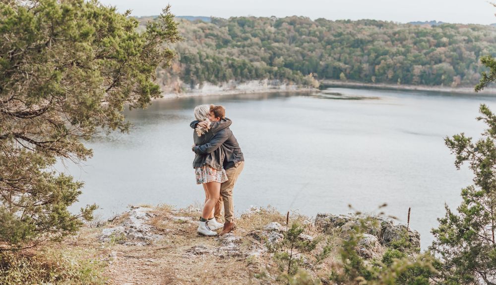 A couple embraces while standing on a rocky cliff overlooking a scenic lake surrounded by forested hills.