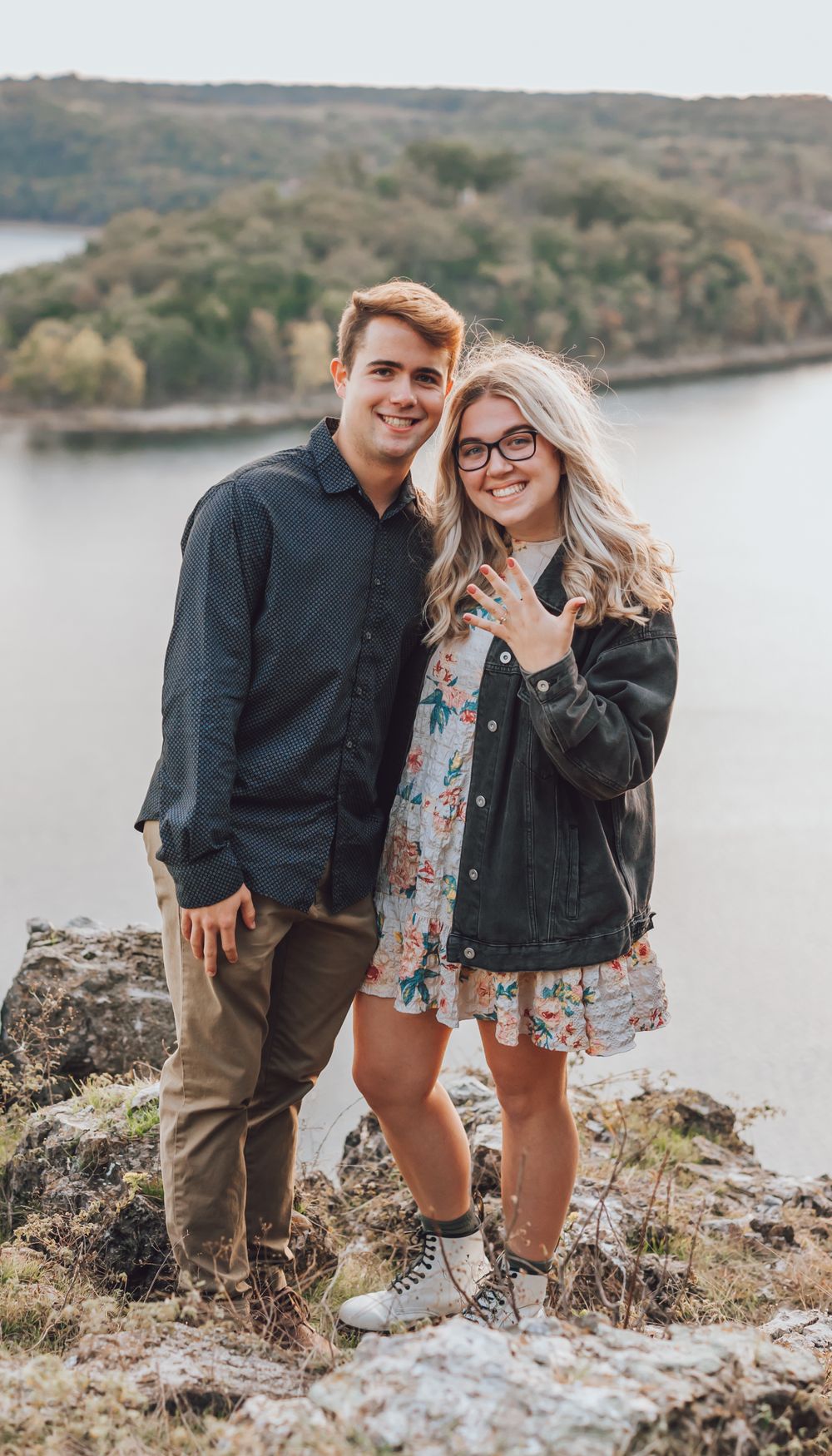 A couple poses together on a rocky cliff overlooking a scenic lake on an autumn day.