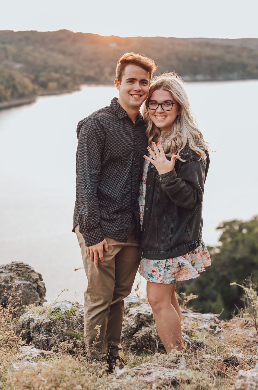A couple stands together on a rocky overlook during sunset at a scenic lake surrounded by hills.