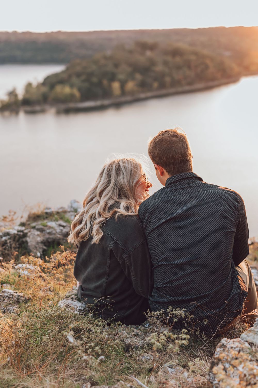 Couple sitting together on rocky cliff overlooking serene lake at sunset during autumn.