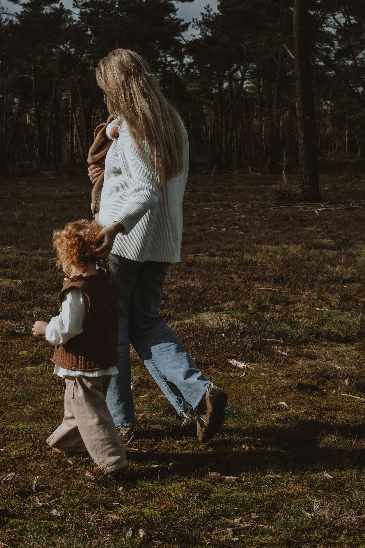 Mother walking with young curly-haired child through a sunlit forest clearing, holding hands.