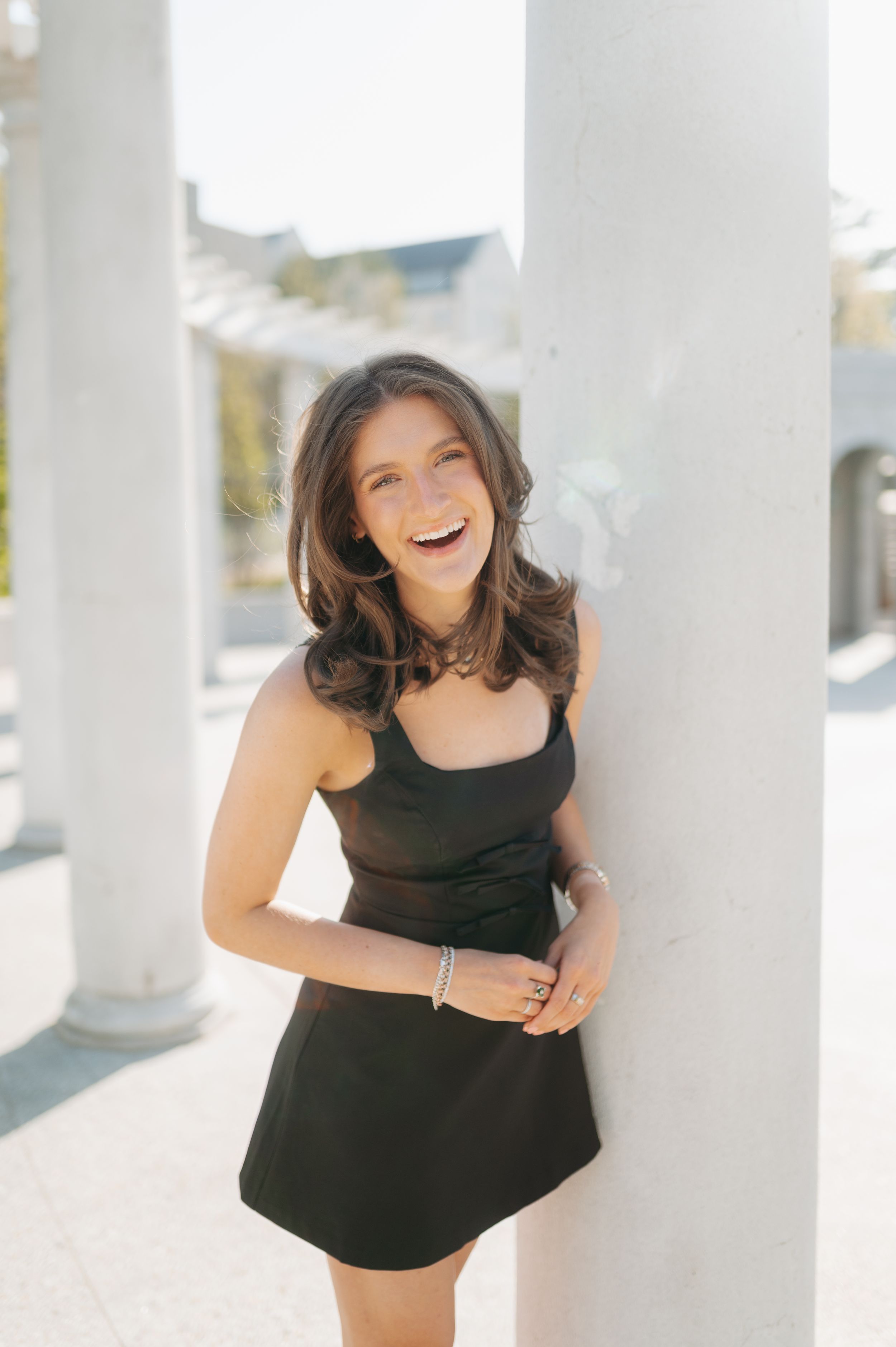 Laughing brunette woman in black mini dress leaning against white column in bright outdoor setting.