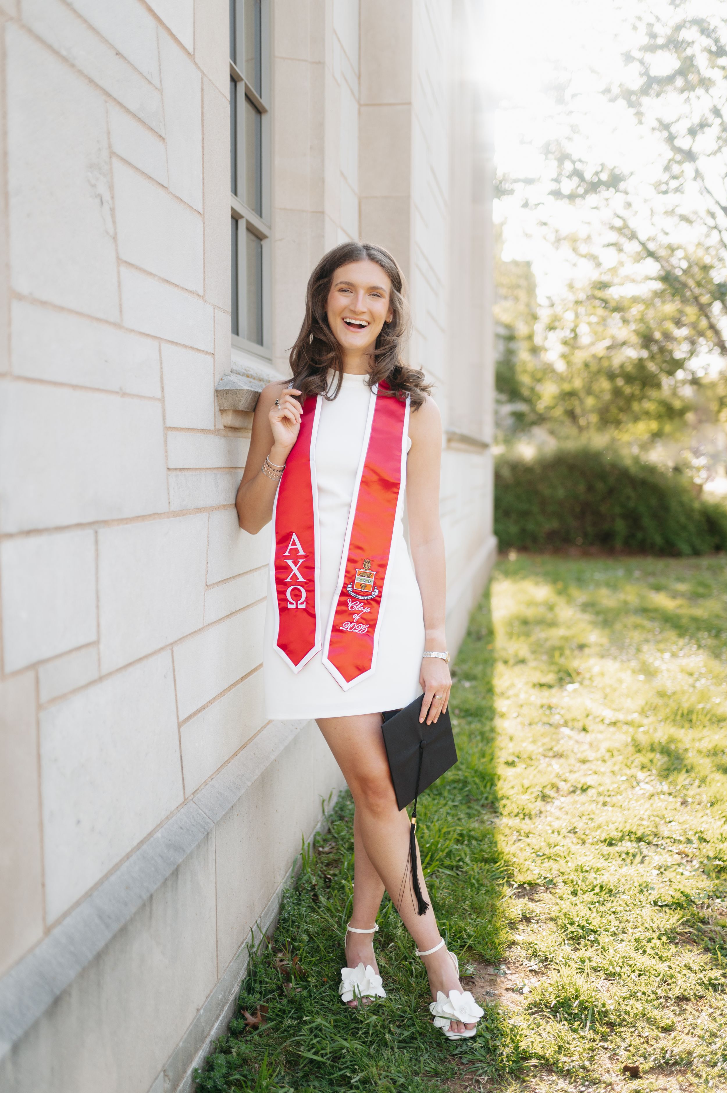 Smiling graduate in white dress wearing red Alpha Chi Omega sorority stole leaning against stone wall.