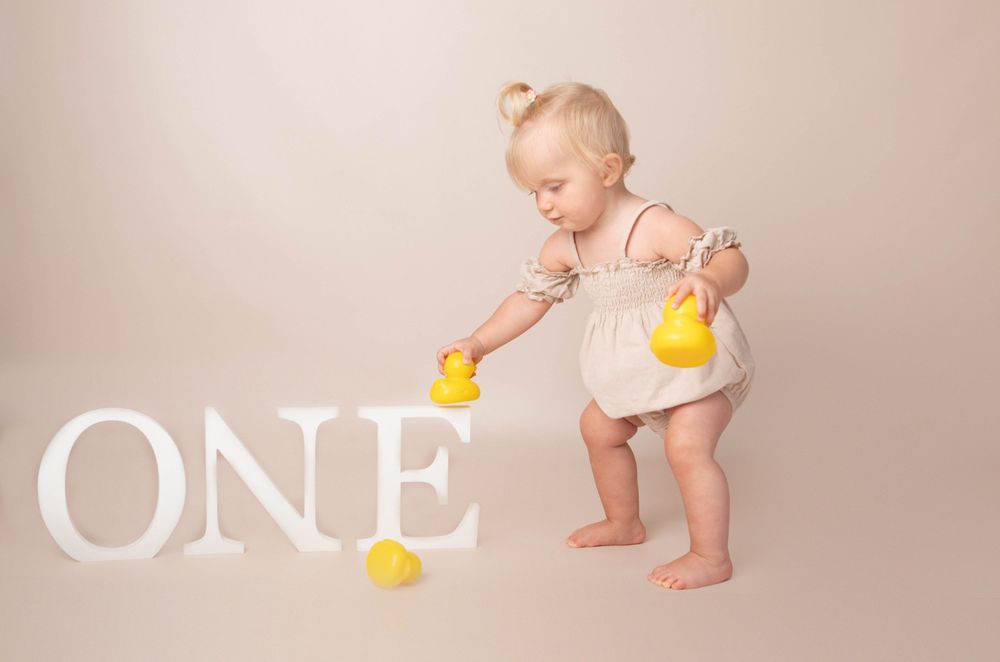 Cute toddler girl in beige dress playing with yellow rubber ducks near white ONE letters.