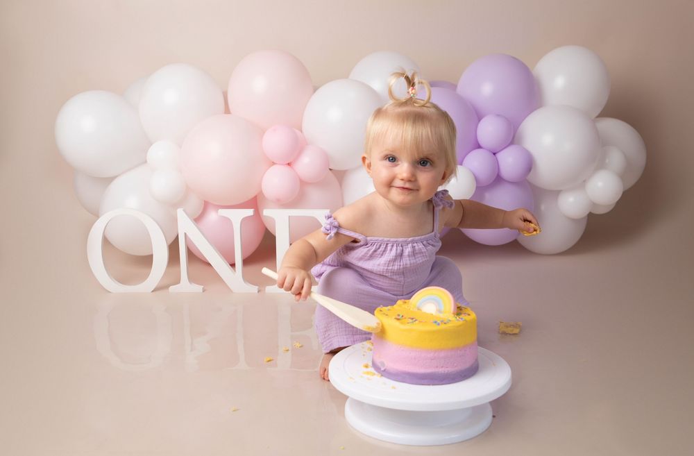 Smiling baby girl in purple dress with first birthday cake smash setup and balloon garland backdrop.