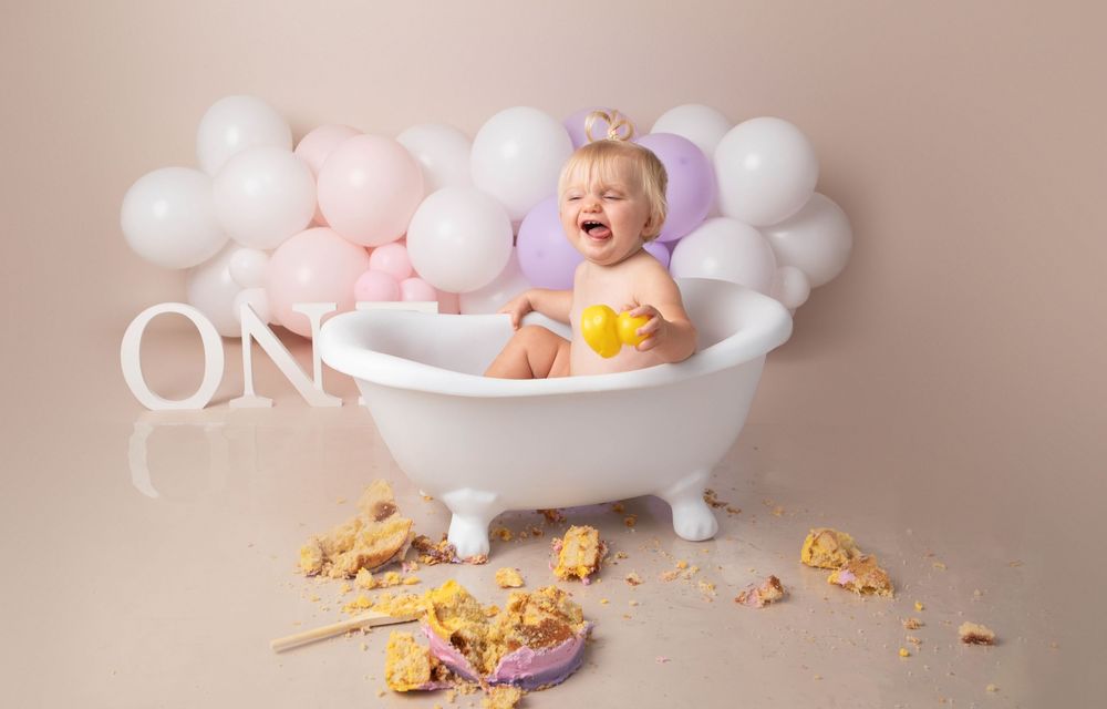 Baby girl laughing in white clawfoot bathtub with pink balloons and smashed cake pieces around her.