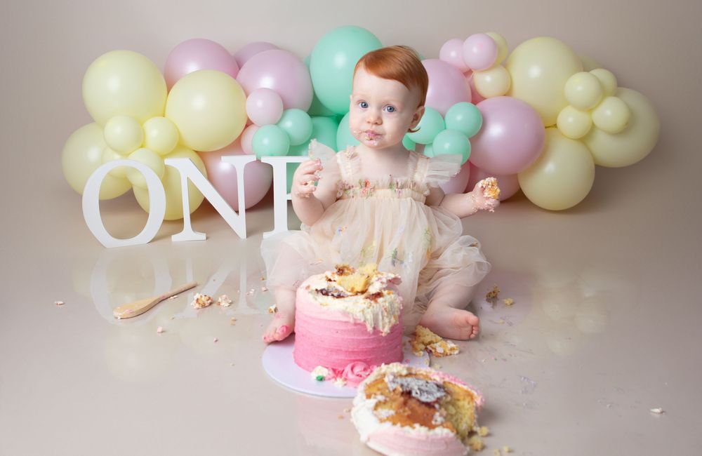 Baby girl in tutu dress smashing first birthday cake surrounded by pastel balloons and ONE sign.