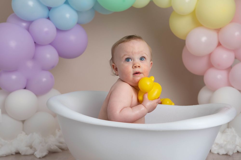 Blue-eyed baby holds yellow rubber duck in white bathtub surrounded by pastel rainbow balloon arch.