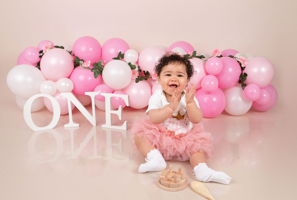 Happy baby girl in pink tutu clapping hands at first birthday cake smash with pink balloon backdrop.