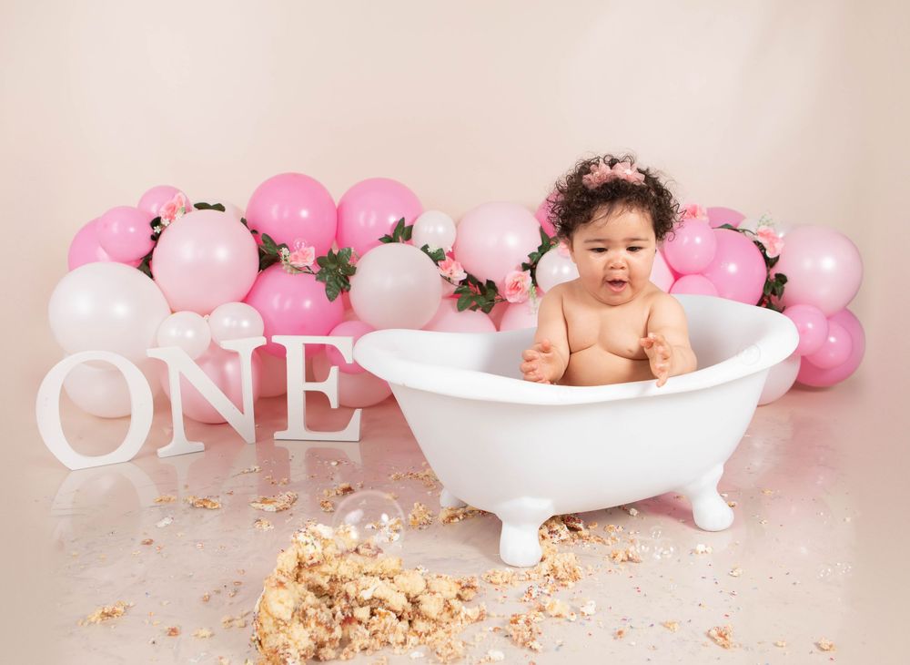 Baby girl sitting in white clawfoot tub with pink balloon garland and smashed cake nearby.