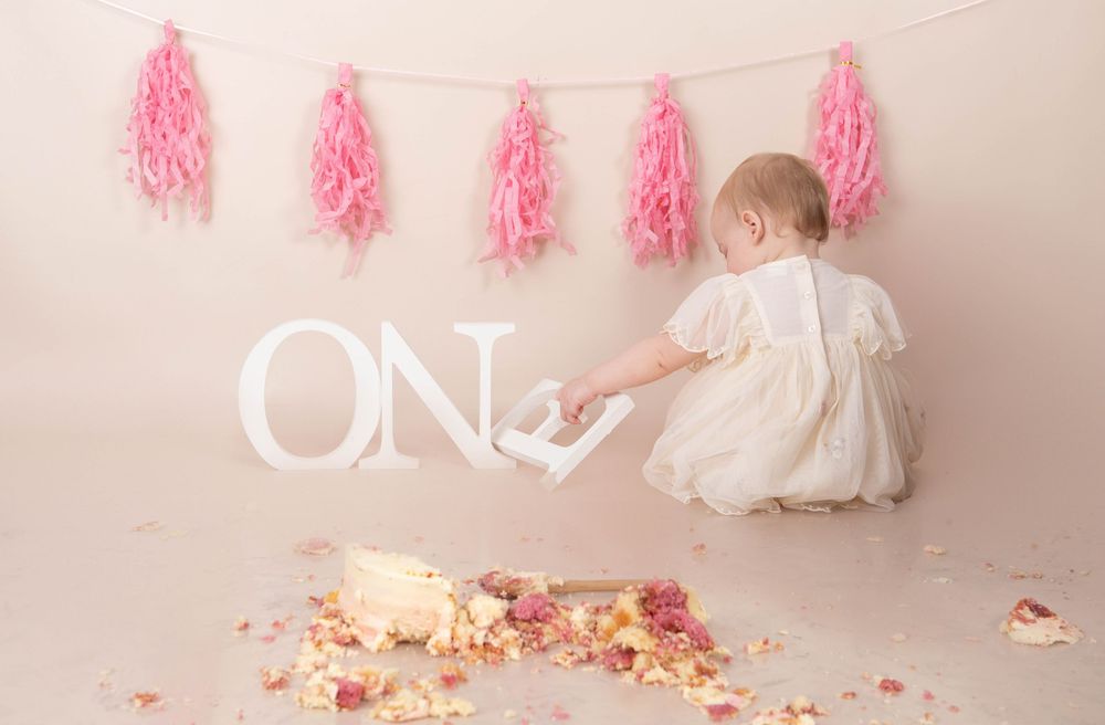 Baby in white dress reaching for ONE letters with smashed floral cake and pink tassels backdrop.