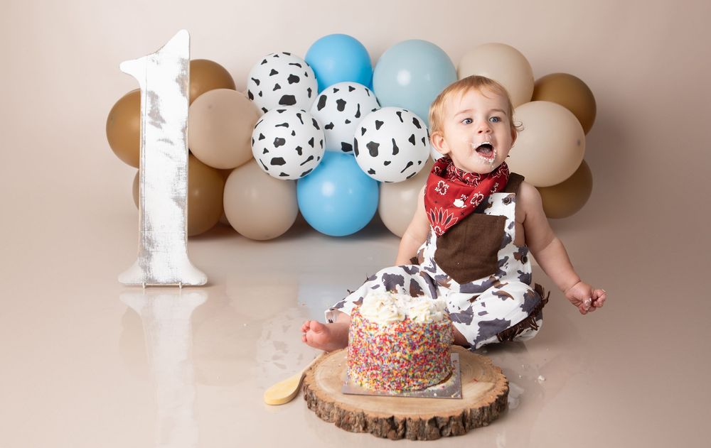 Baby boy in cowboy outfit celebrating first birthday with cake smash and balloon backdrop.