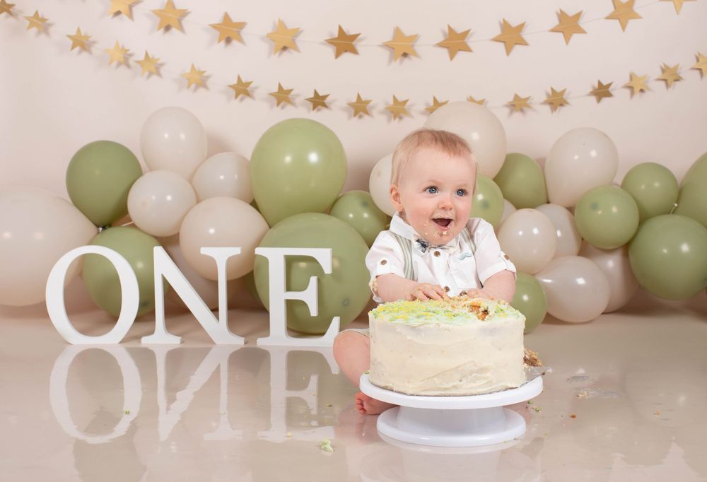 Happy baby smashes first birthday cake surrounded by green and white balloons and ONE sign.