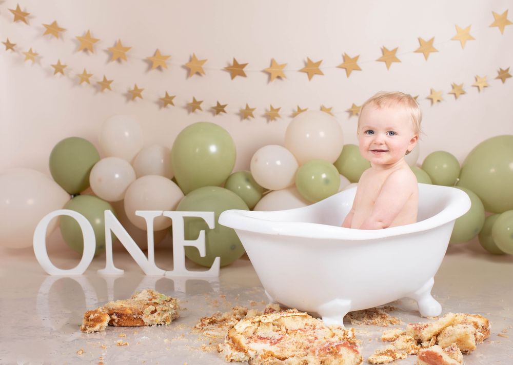 Smiling baby sits in white bathtub with smashed cake and green balloons for first birthday.