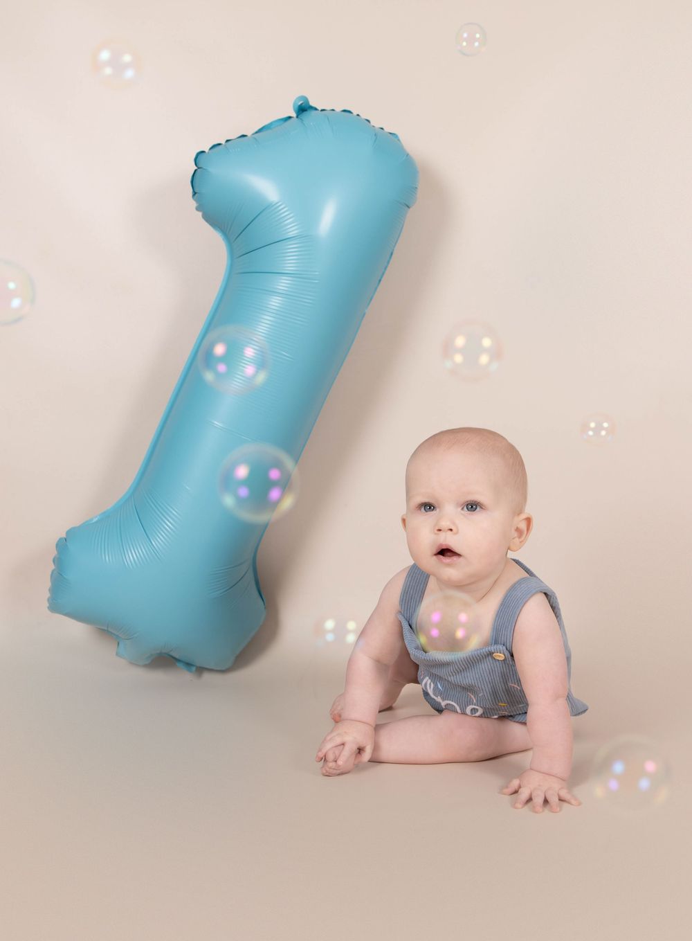 Curious baby boy sitting beside large blue number one balloon with soap bubbles floating around.
