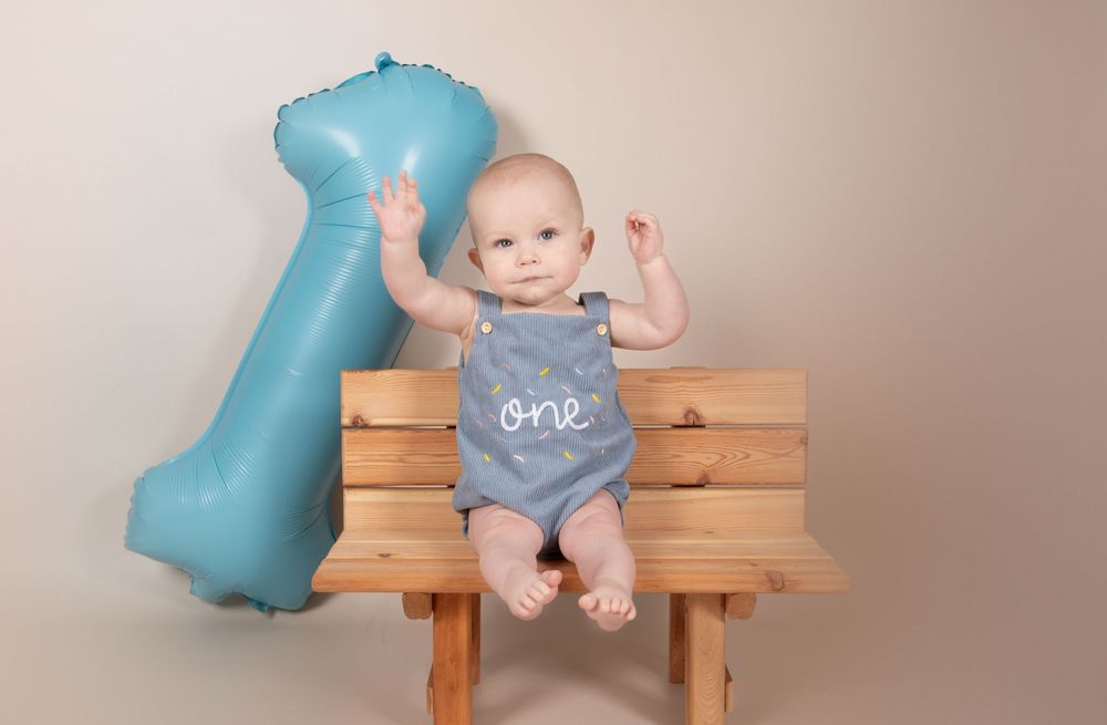Baby in blue 'one' romper sits on wooden bench beside blue number one balloon for first birthday photo.