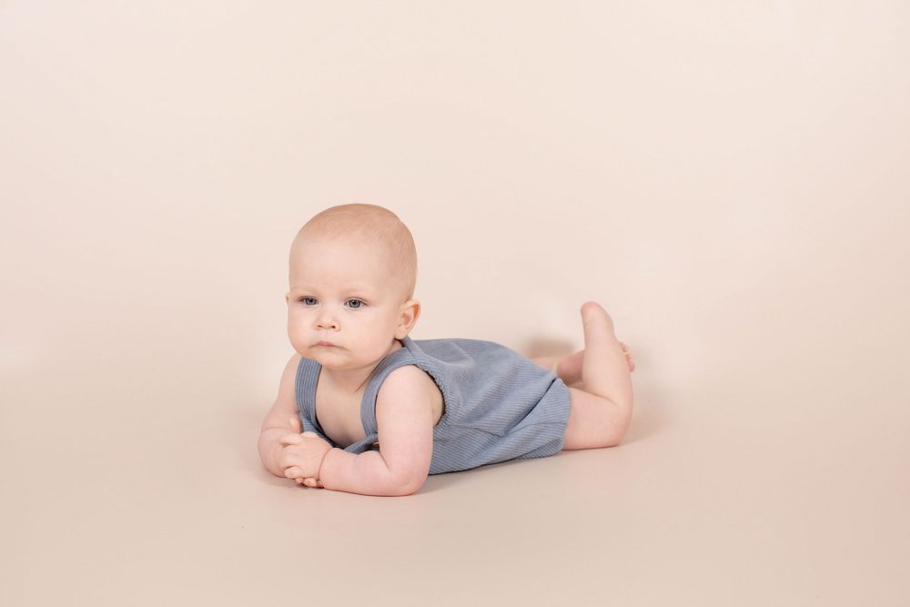 Adorable baby in blue romper doing tummy time on a neutral beige studio background.