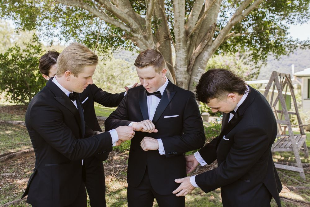 Groomsmen in black tuxedos help each other with final preparations outdoors.