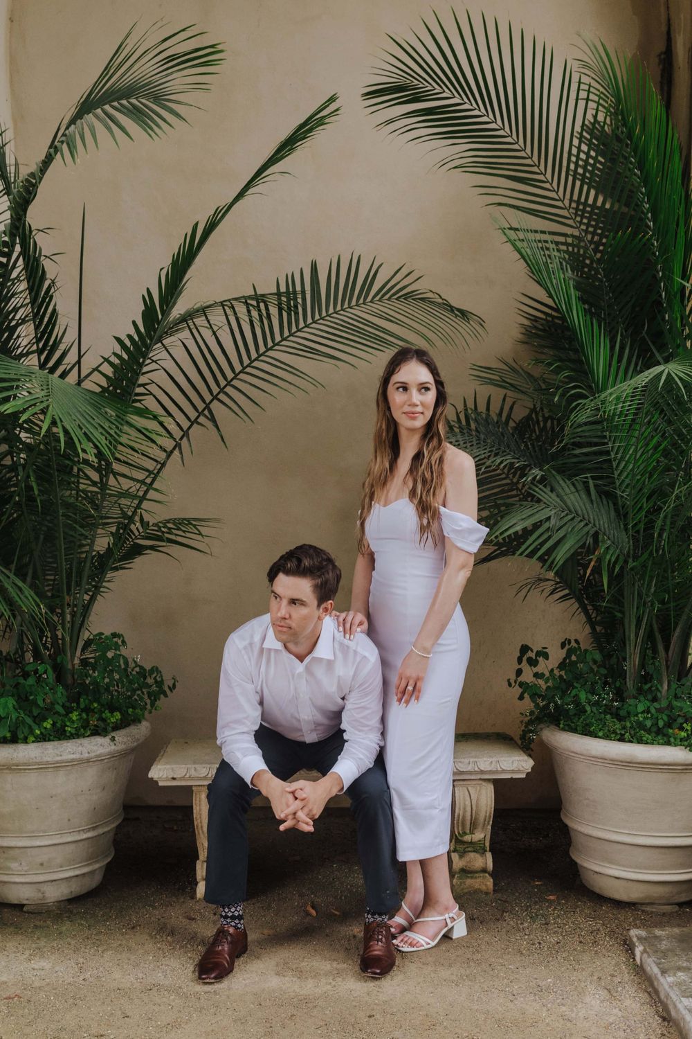 Stylish couple poses with lush tropical palm plants in decorative pots against a neutral beige wall.