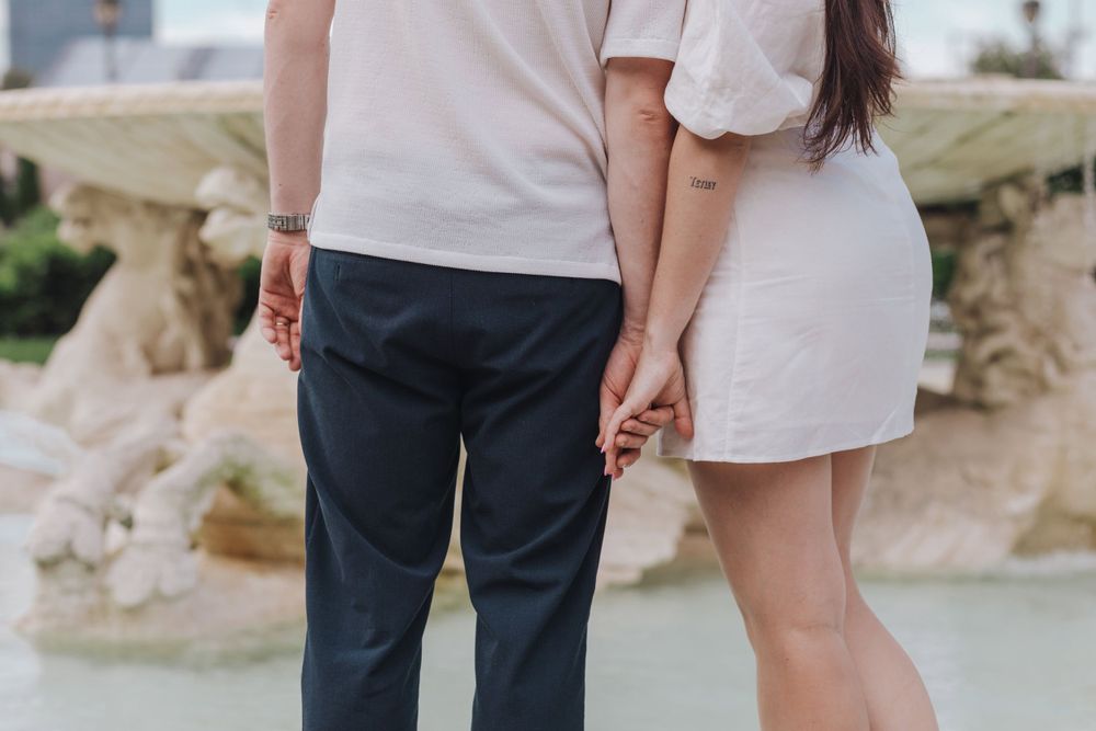 A couple in light colored clothing hold hands while standing together on a classical stone terrace.