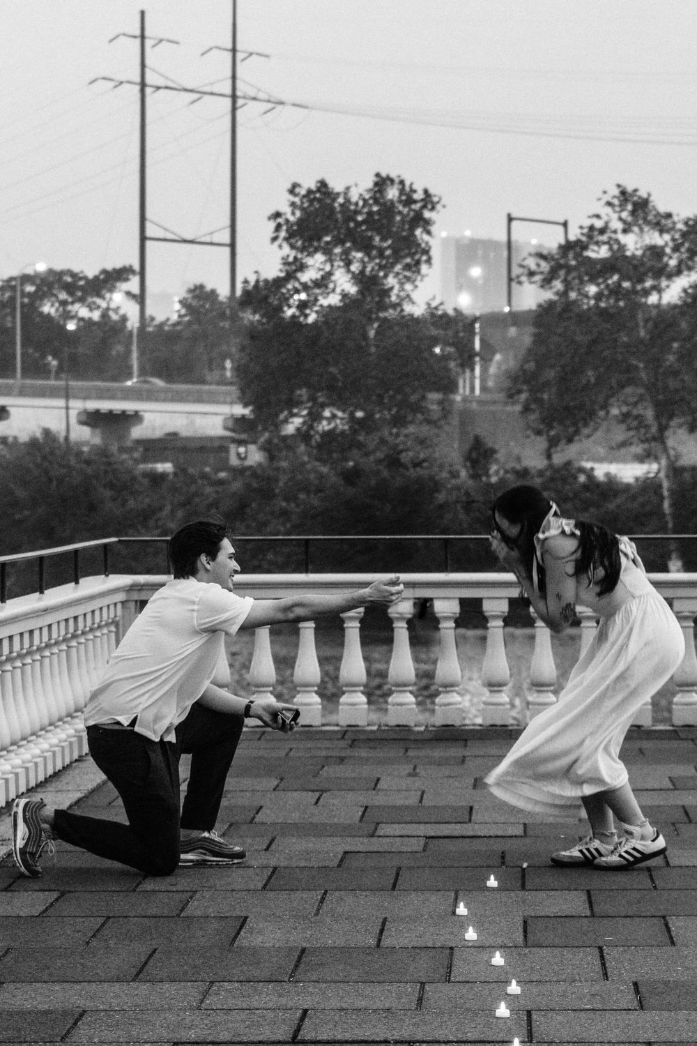 A candlelit proposal scene under a stone balustrade at dusk with power lines visible in background.