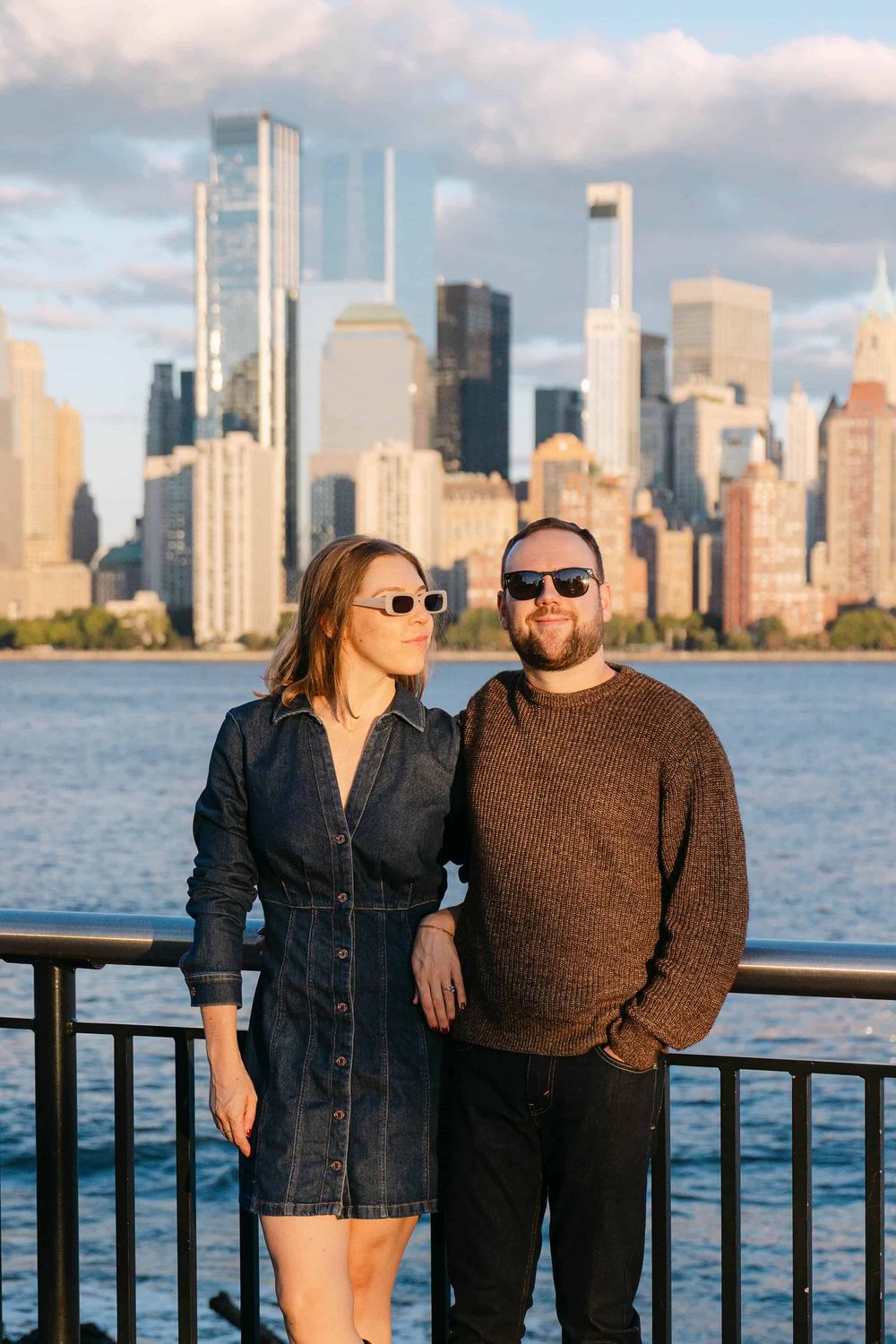 A couple poses along the waterfront with the Manhattan skyline visible across the water during daytime.