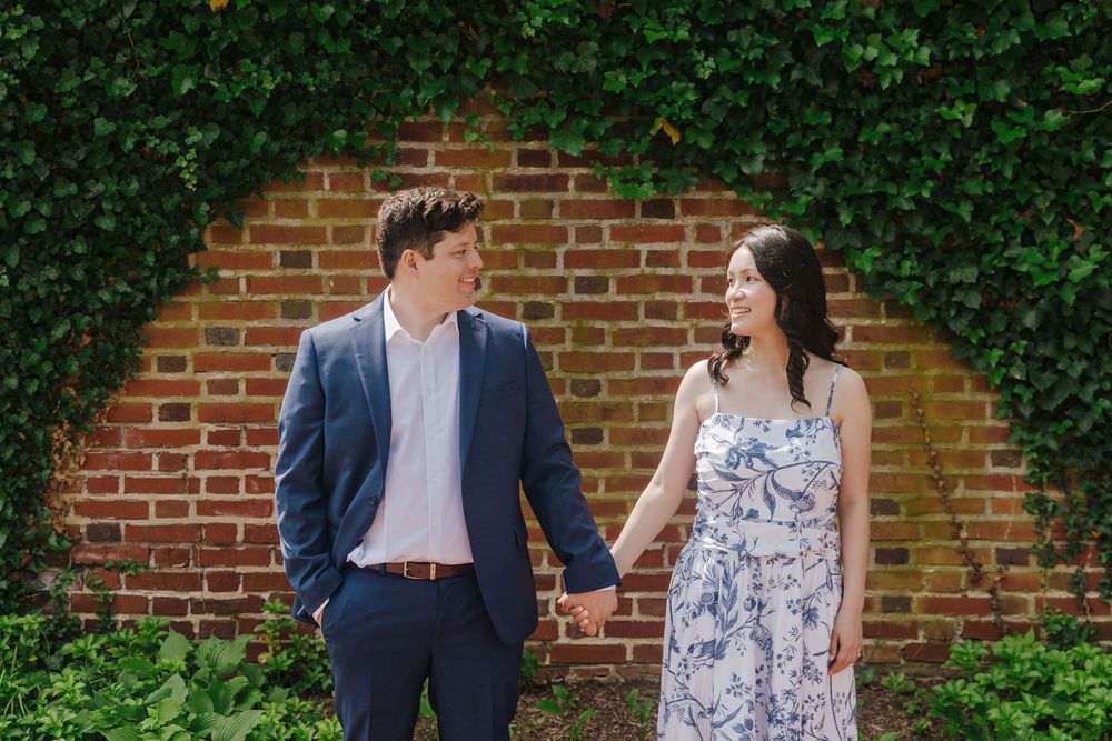 Couple stands together smiling by an ivy-covered brick wall in a garden setting.