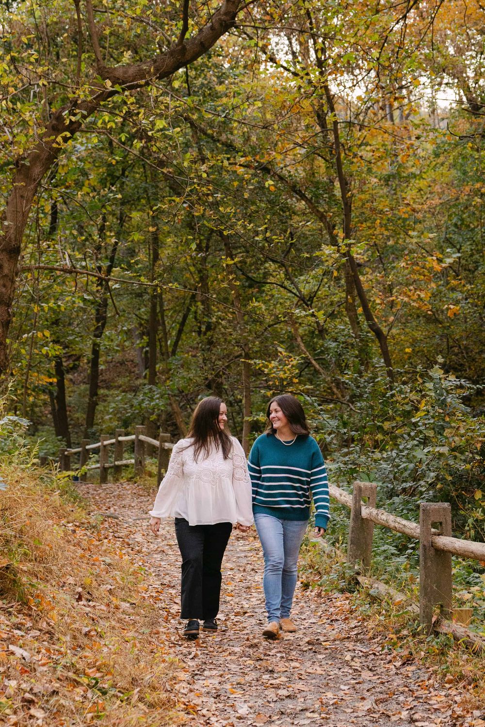 Two people stroll down a leaf-covered forest path surrounded by trees with autumn colors.
