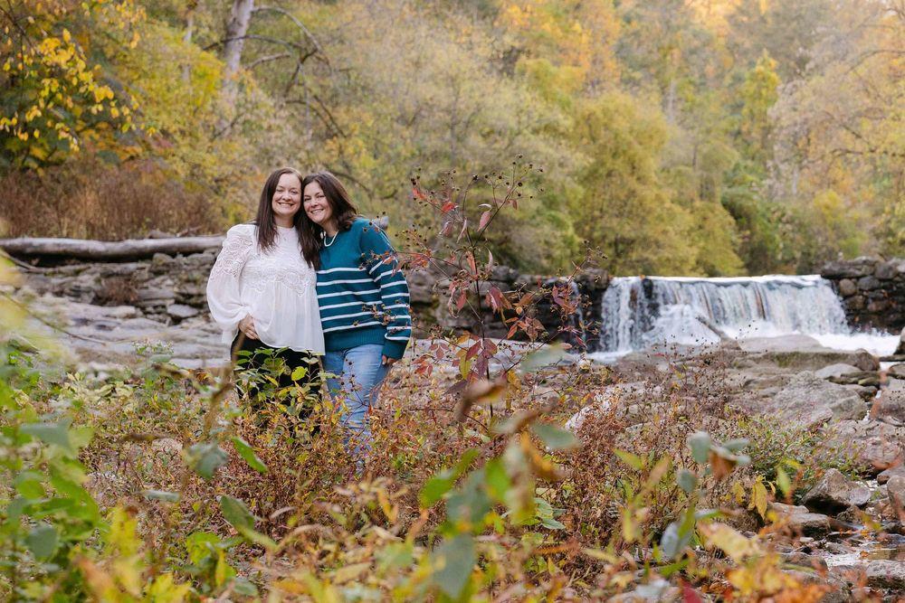 A couple walks along an autumn trail with a waterfall and colorful fall foliage in the background.