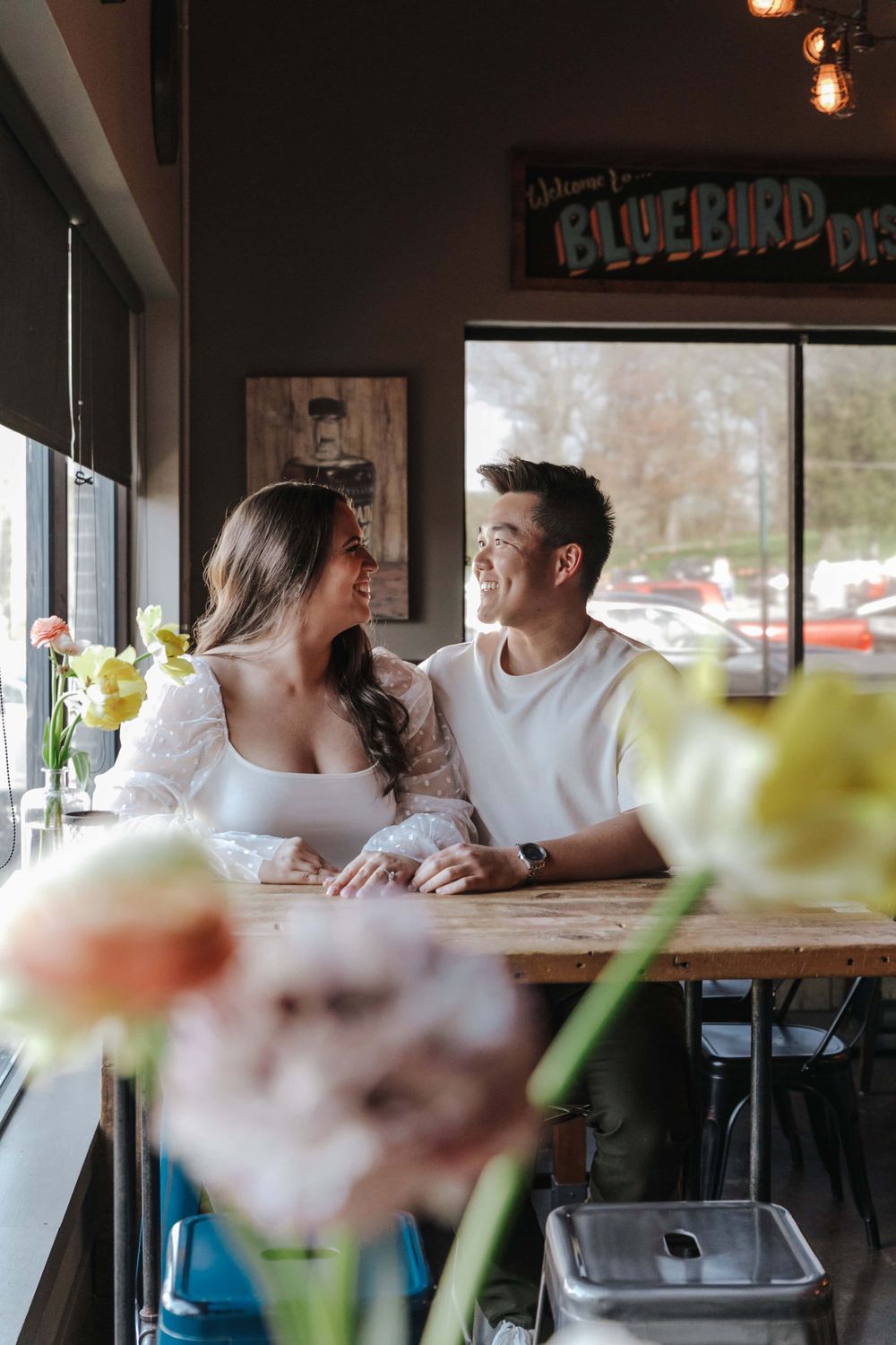 A couple shares an intimate moment while sitting at a rustic restaurant table with flowers in the foreground.