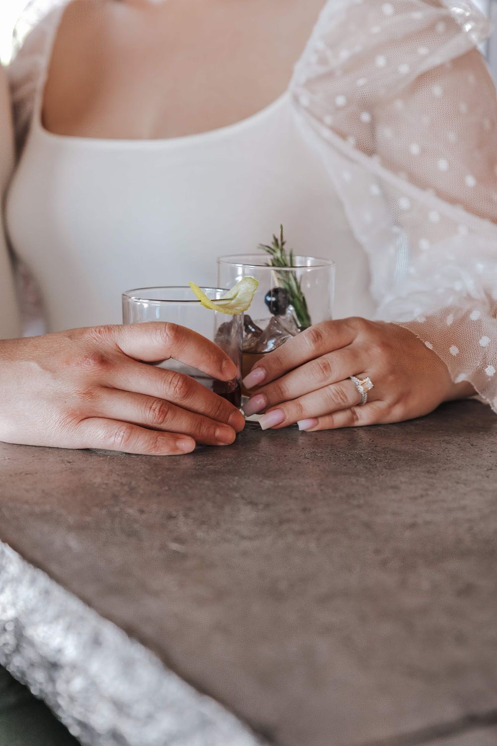 Elegant hands wearing a wedding ring hold a delicate cocktail glass with citrus and herbs on a textured surface.