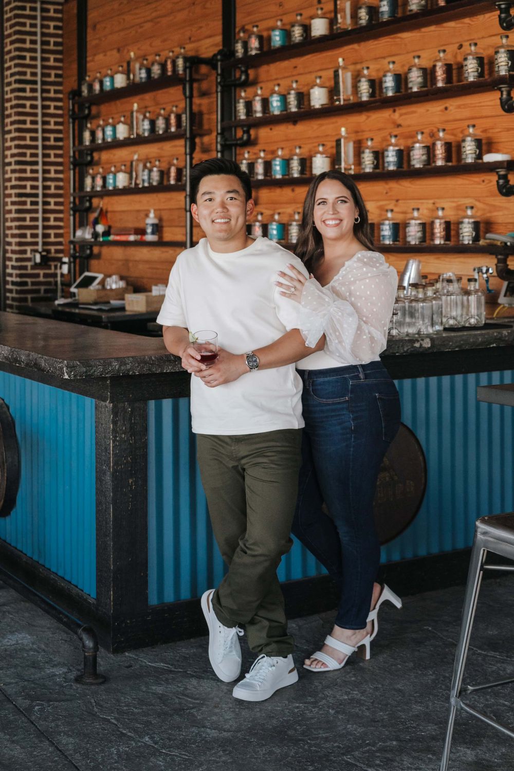 Two people lean against a blue bar counter with wooden walls lined with glass jars and bottles in the background.