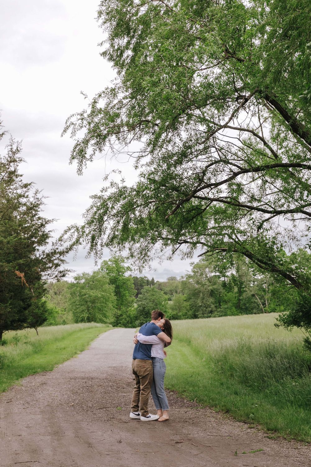 A couple shares an intimate moment on a tree-lined path through a lush green park.