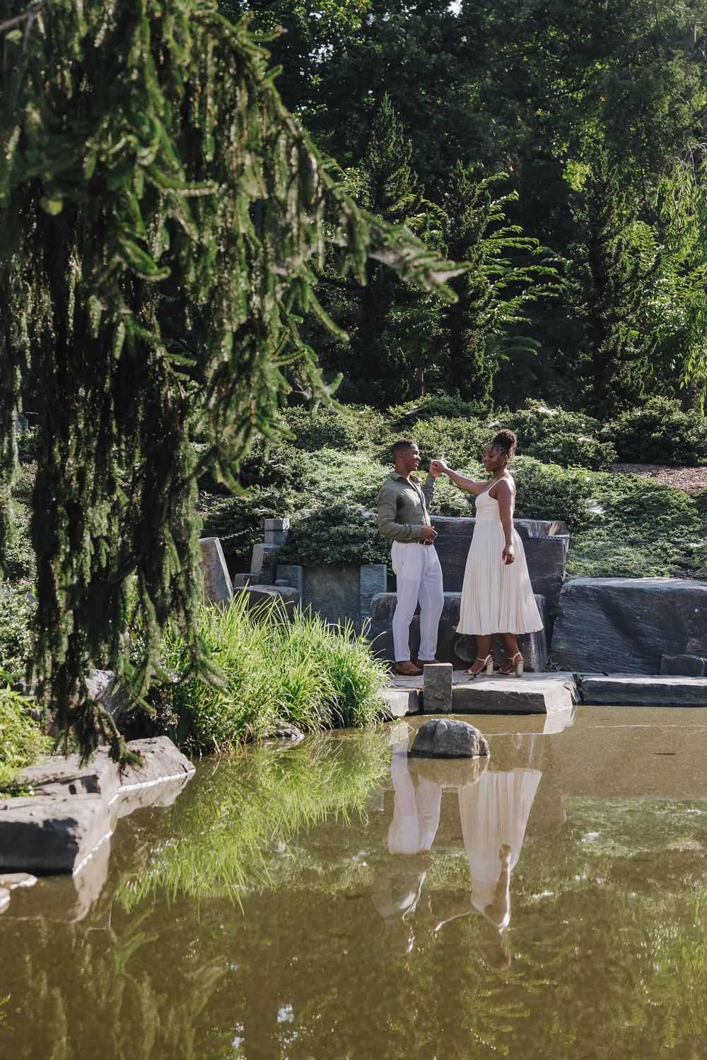 Two people in white outfits stand near a serene pond surrounded by lush greenery in a garden setting.