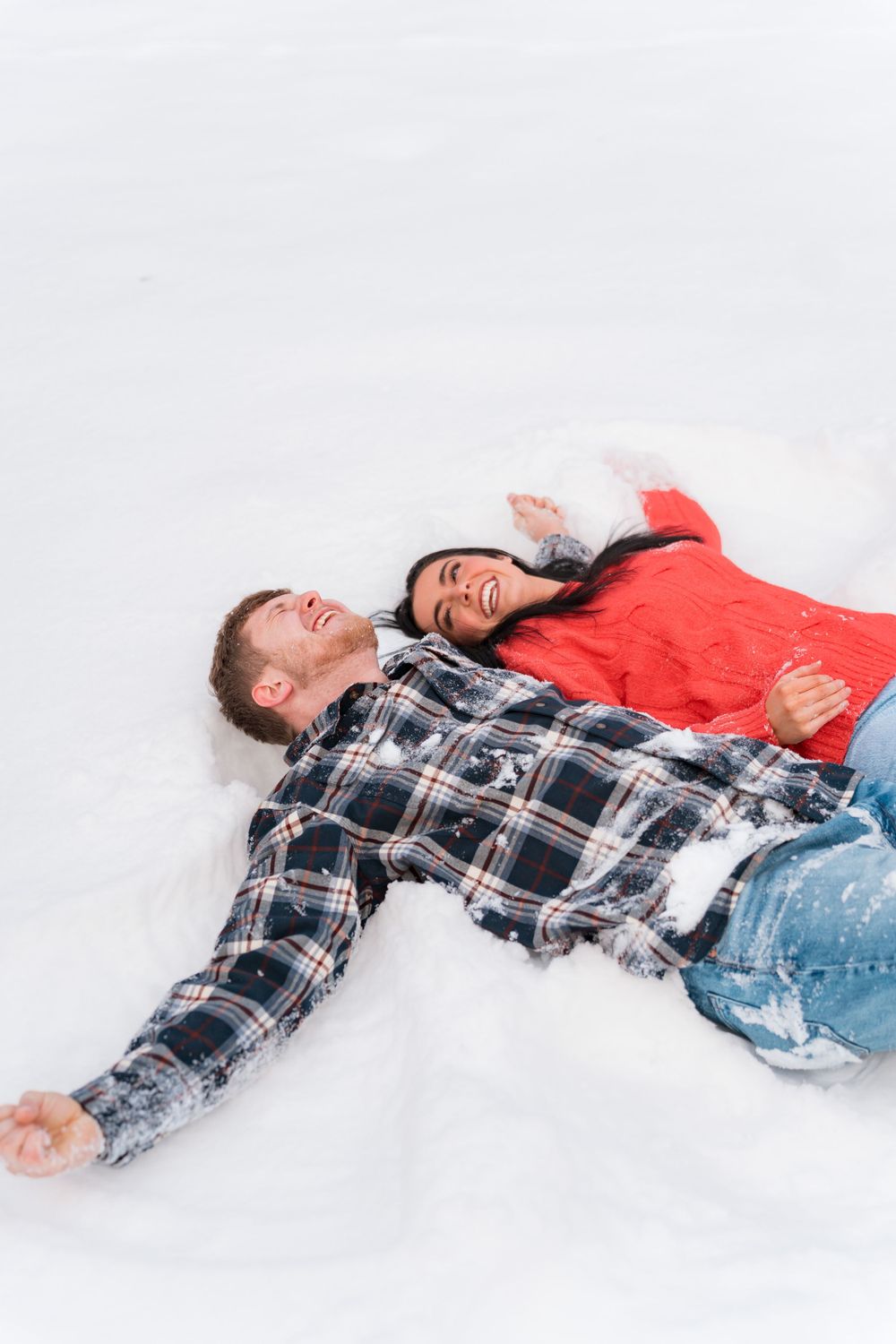 Two people lay in the snow making snow angels while wearing winter clothes and laughing together.