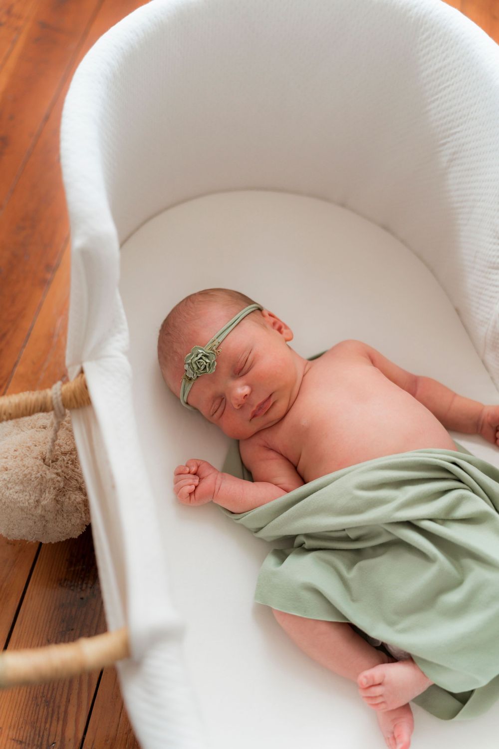 A sleeping newborn wrapped in sage green fabric lies peacefully in a white bassinet on wooden floor.