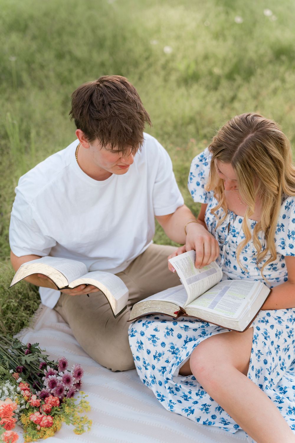 Two people sit on a blanket reading books together outdoors with flowers nearby.