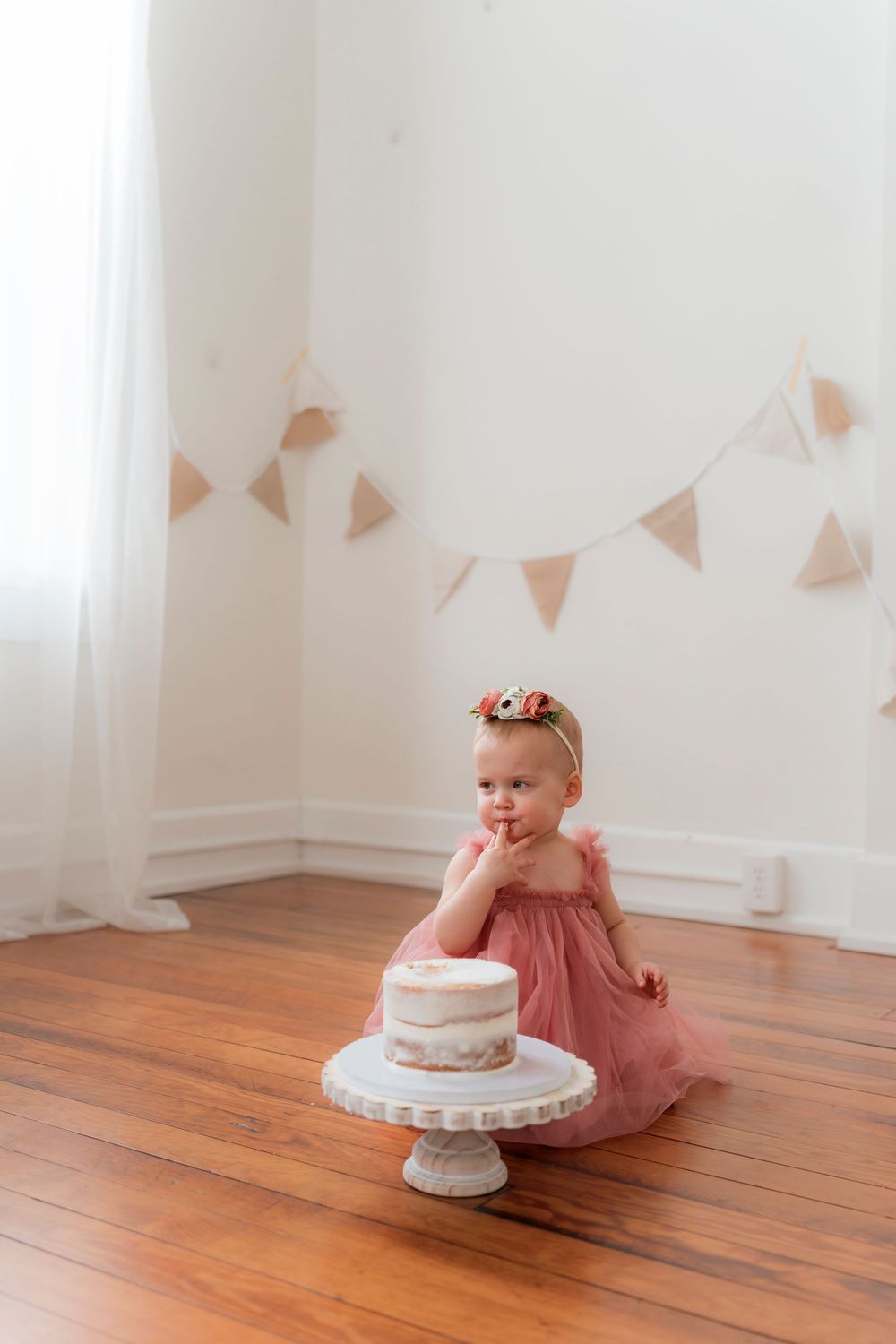 Small child in pink dress sits on hardwood floor beside white cake on pedestal with bunting decorations above.