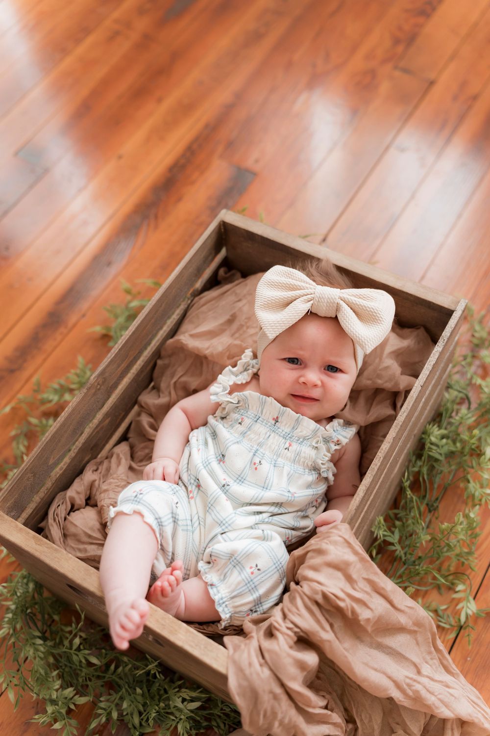 Baby wearing white patterned romper and bow lies in wooden box with brown fabric and greenery decorations on hardwood floor.