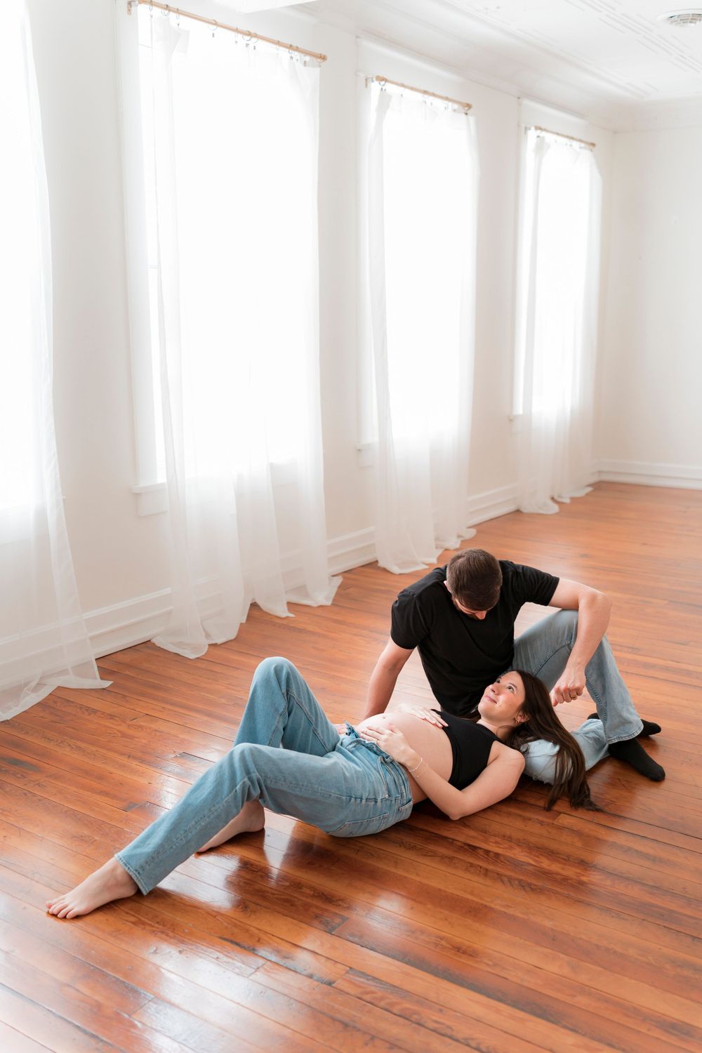 Couple laying together on polished hardwood floor beside sheer white curtained windows in bright minimalist room.