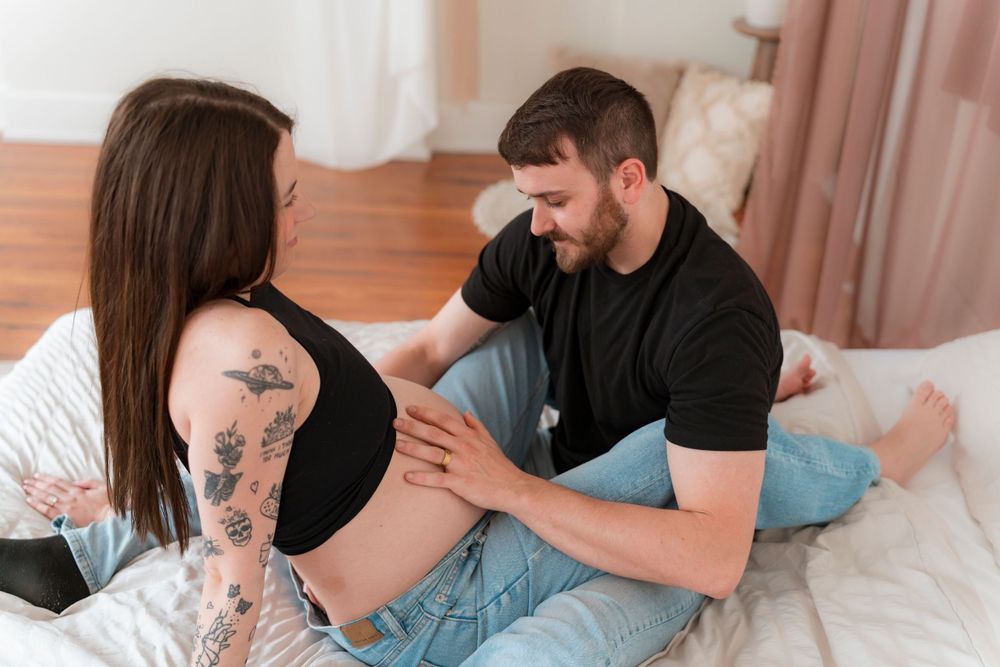 Couple sharing intimate moment on bed with woman showing visible arm tattoos in casual home setting.