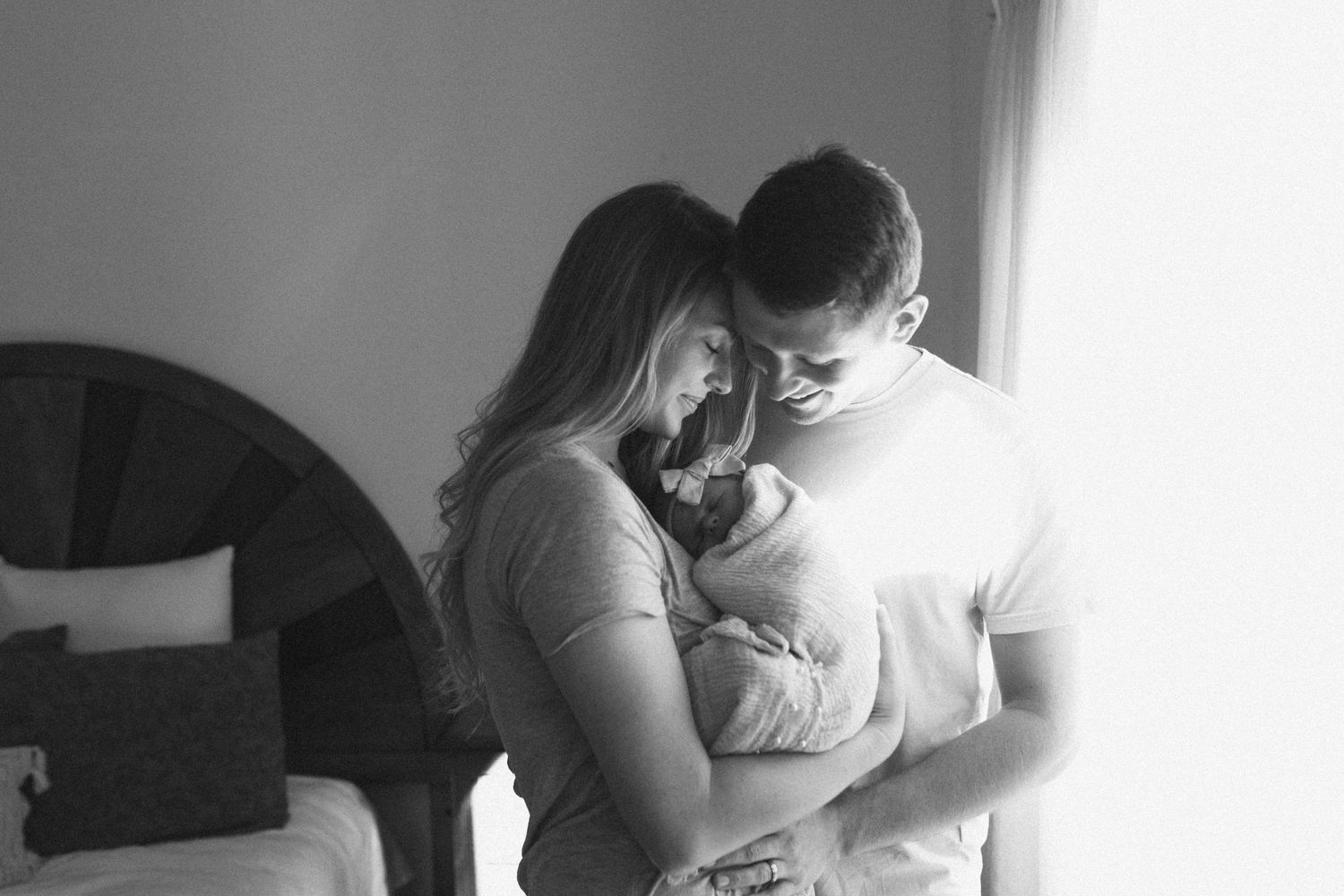 Photo by B.Logsdon of a tender moment in black and white as two people gaze down at their newborn baby together in soft window light.