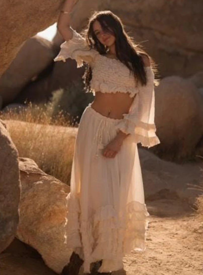 A woman in a bohemian white two-piece outfit poses in golden sunlight against desert rocks and dry grass.